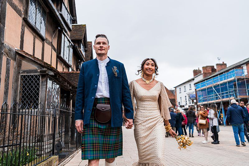 A couple walks hand-in-hand in festive attire; the man wears a kilt and blue jacket, and the woman wears a patterned dress, holding a bouquet. They are on a street with historic buildings and pedestrians. Image by Documentary Wedding Photographer Lewis Bishop Photography.