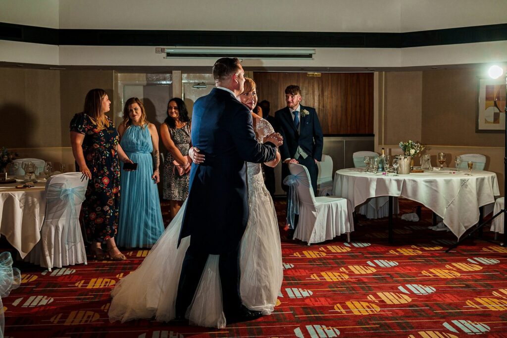 A bride and groom share their first dance at a wedding reception, with the bride in a white gown and the groom in a dark suit. Guests in elegant attire watch and smile in the background, standing around decorated tables in a dimly lit room. Image by Documentary Wedding Photographer Lewis Bishop Photography.