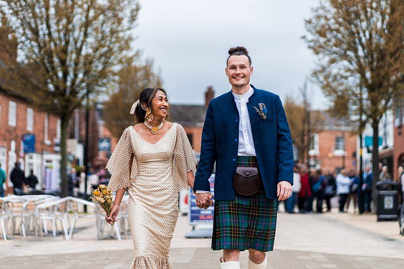A couple dressed in wedding attire, with the man in a tartan kilt and the woman in a beige dress, walk hand in hand down a tree-lined street. Image by Documentary Wedding Photographer Lewis Bishop Photography.