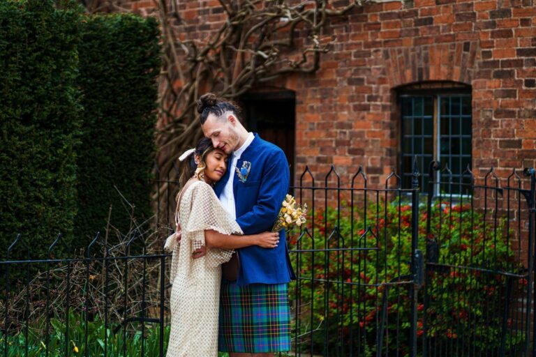 A couple embraces outdoors, with the man in a blue jacket and tartan kilt, and the woman in a patterned dress holding a golden bouquet. They stand in front of a brick building and a black fence. Image by Documentary Wedding Photographer Lewis Bishop Photography.