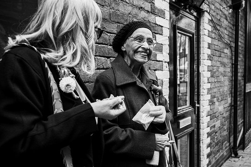 Two women stand outside a brick building, engaged in conversation. One woman, wearing glasses and a beanie, smiles while holding a paper and a bag. The other woman holds a small flower. Image by Documentary Wedding Photographer Lewis Bishop Photography.