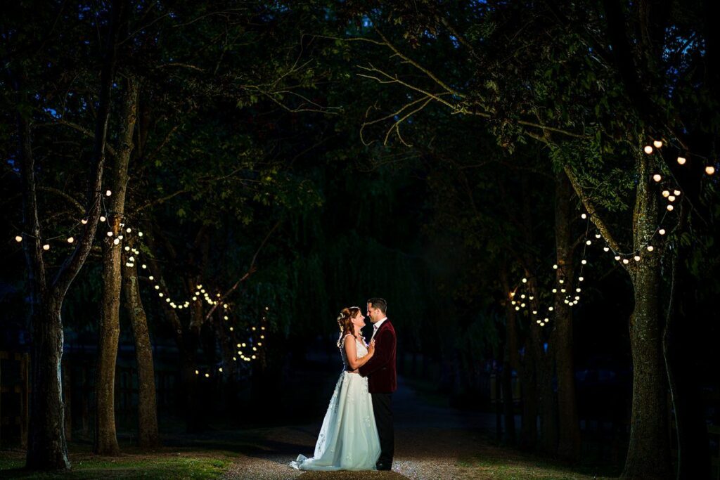 A newlywed couple shares a romantic moment under a canopy of trees adorned with string lights at night. The bride wears a flowing white gown, and the groom is dressed in a dark suit as they embrace, illuminated by the warm light. Image by Documentary Wedding Photographer Lewis Bishop Photography.
