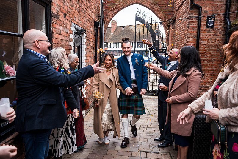 A newlywed couple walks through an archway lined with smiling and clapping guests. The groom is wearing a kilt, and the bride is in a long dress, holding a bouquet. Image by Documentary Wedding Photographer Lewis Bishop Photography.