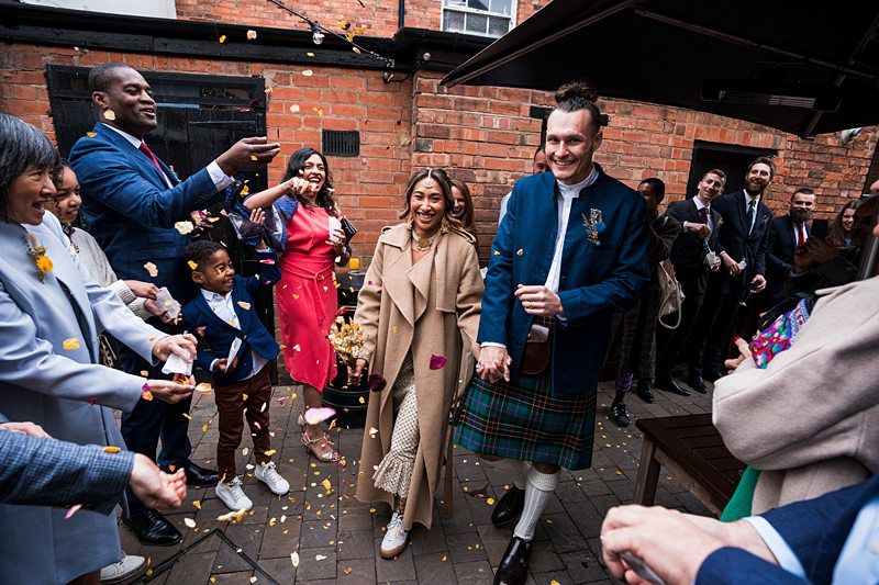 A bride in a beige coat and groom in a kilt walk through a crowd throwing petals in an outdoor venue with brick walls. Image by Documentary Wedding Photographer Lewis Bishop Photography.