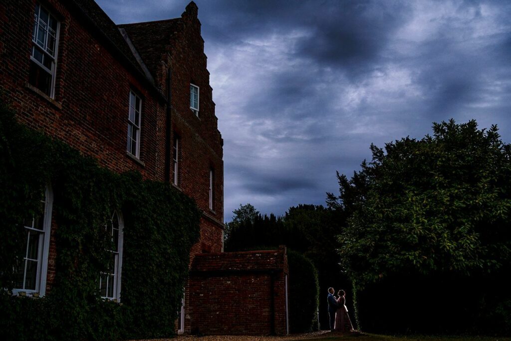 A couple stands close together in a tender moment outside a large, ivy-covered brick building during evening twilight. The sky above is filled with dark, dramatic clouds, adding an atmospheric backdrop to the intimate scene. Image by Documentary Wedding Photographer Lewis Bishop Photography.