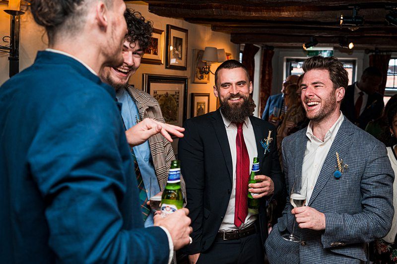 A group of four men in suits are standing and talking while holding drinks at a social gathering. They appear to be enjoying themselves and laughing. Image by Documentary Wedding Photographer Lewis Bishop Photography.