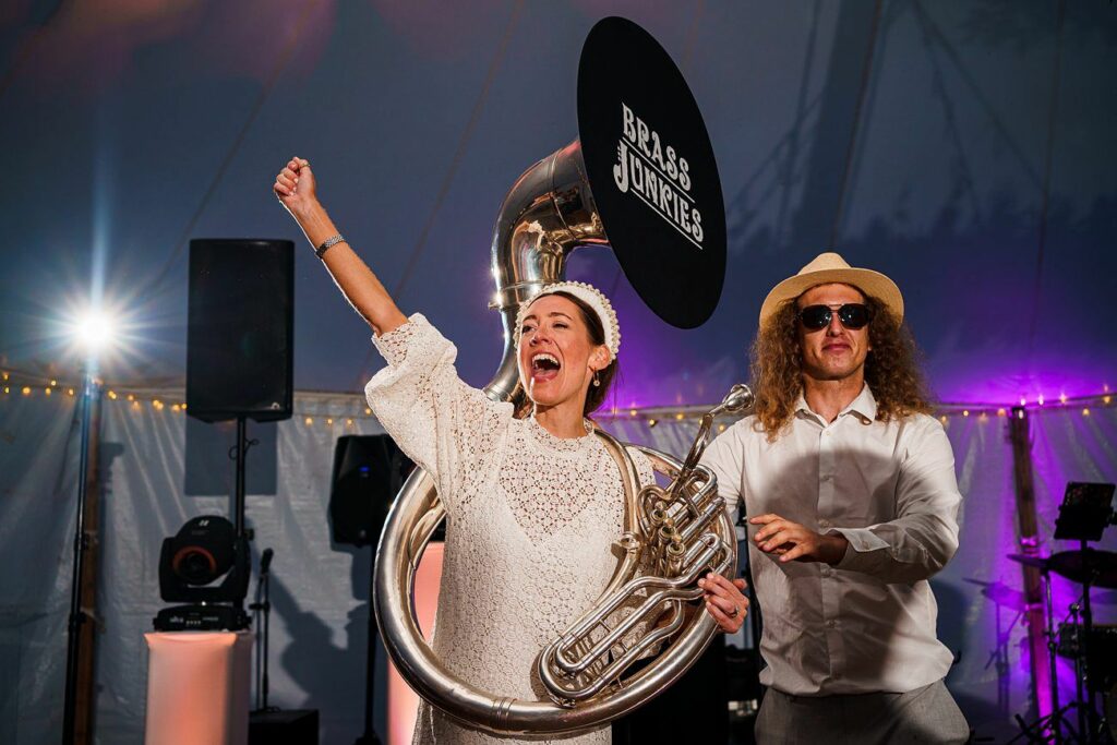 A joyous woman in a white dress energetically plays a large brass instrument, her arm raised. A man in sunglasses and a hat stands beside her, joining in the celebration. The background is illuminated with colorful stage lights and fairy lights. A "BRASS JUNKIES" sign is visible. Image by Documentary Wedding Photographer Lewis Bishop Photography.