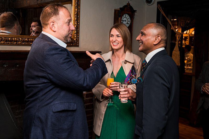 Three people are conversing and smiling at a social gathering. One man is gesturing while speaking, and the other man and woman are holding drinks. Image by Documentary Wedding Photographer Lewis Bishop Photography.