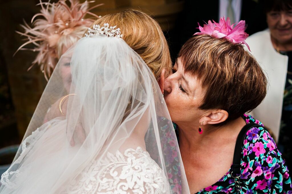A woman in a colorful floral dress and pink fascinator kisses the cheek of a bride wearing a white wedding dress with lace details and a veil adorned with a tiara. Another woman wearing a beige outfit with a pink fascinator stands close by and watches. Image by Documentary Wedding Photographer Lewis Bishop Photography.