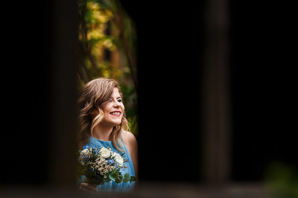 A woman with wavy blonde hair, wearing a light blue dress, is smiling and holding a bouquet of white and blue flowers. She stands in a dimly lit area with green foliage in the background. The image is framed by dark, blurry foreground elements. Image by Documentary Wedding Photographer Lewis Bishop Photography.
