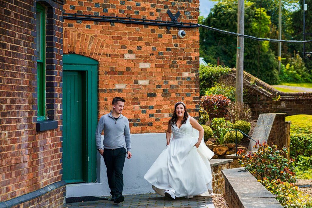 A woman in a white wedding dress and a man in a grey sweater smile while walking outside near a brick building with a green door. Image by Documentary Wedding Photographer Lewis Bishop Photography.