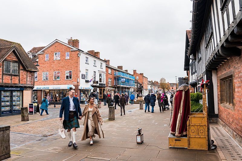People stroll through a busy street with shops and buildings, including a performer in a historical outfit standing by an ornate box. Image by Documentary Wedding Photographer Lewis Bishop Photography.