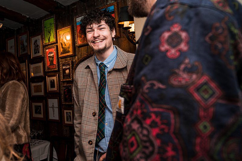 A man in a brown checkered jacket and tie smiles at a social gathering, standing near another person who is partially visible in a patterned garment. Artwork hangs on the wall in the background. Image by Documentary Wedding Photographer Lewis Bishop Photography.