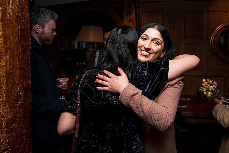 Two women hugging in a warmly lit room with a wooden interior. In the background, a man converses with another person holding a drink. Image by Documentary Wedding Photographer Lewis Bishop Photography.