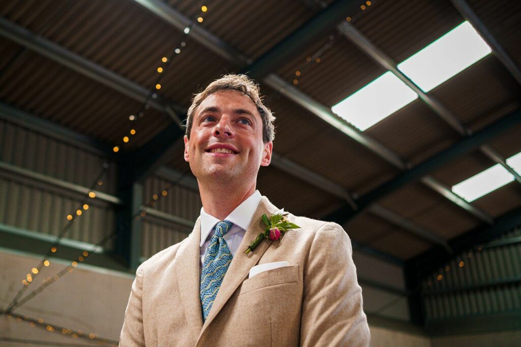 A man wearing a beige suit and blue patterned tie stands indoors, smiling. He has a boutonniere on his left lapel. The background shows a high ceiling with exposed beams and string lights. Image by Documentary Wedding Photographer Lewis Bishop Photography.