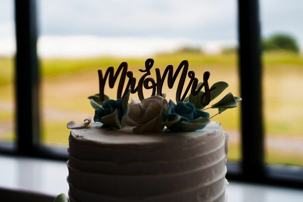 A white frosted cake sits near a window with a scenic view. The cake is topped with a "Mr & Mrs" sign and decorated with blue, green, and white flowers. The background shows a blurred outdoor landscape. Image by Documentary Wedding Photographer Lewis Bishop Photography.