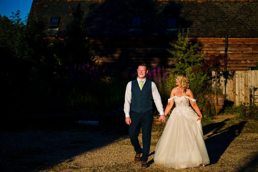 A bride in a white gown and groom in a navy suit walk hand in hand, smiling, in a sunlit outdoor setting with rustic buildings and greenery in the background. The late afternoon sun casts long shadows, illuminating the couple with a warm, golden light. Image by Documentary Wedding Photographer Lewis Bishop Photography.