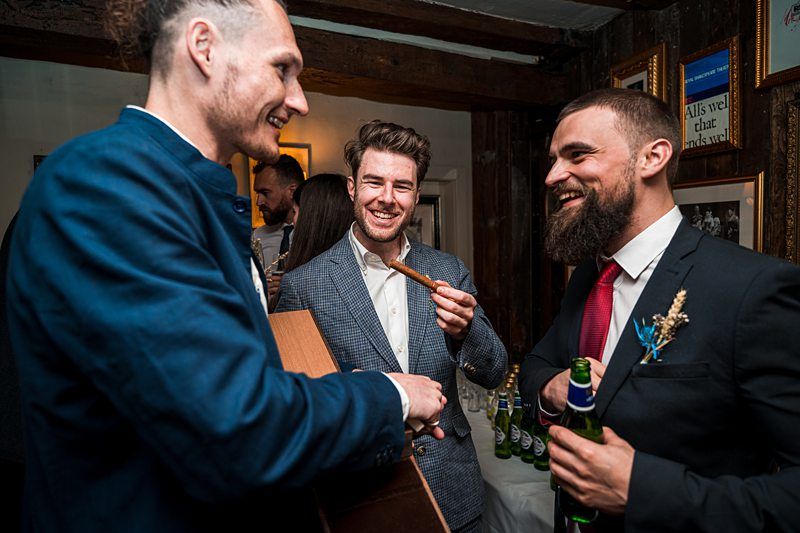 Three men in suits are laughing and enjoying a conversation at a social gathering, with beer bottles and guitar in hand. Framed pictures and decor are visible on the wall behind them. Image by Documentary Wedding Photographer Lewis Bishop Photography.