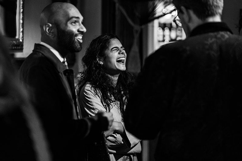 Three people are socializing indoors; one woman is laughing heartily while the two men beside her are engaged in conversation. The photo is in black and white. Image by Documentary Wedding Photographer Lewis Bishop Photography.