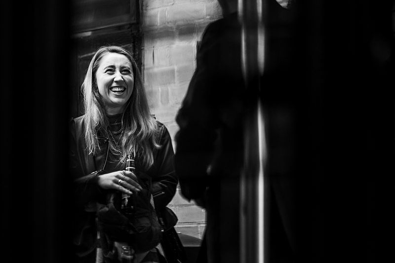 A woman with long hair, holding a drink, smiles while talking to someone in front of her. The image is in black and white. Image by Documentary Wedding Photographer Lewis Bishop Photography.