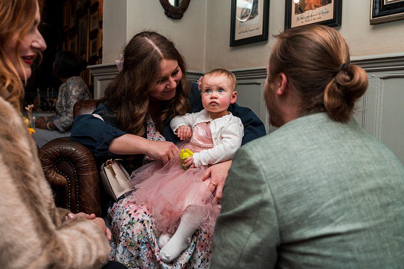 A baby in a pink dress sits on a woman's lap while a man and another woman engage with them in a cozy, decorated room. Image by Documentary Wedding Photographer Lewis Bishop Photography.