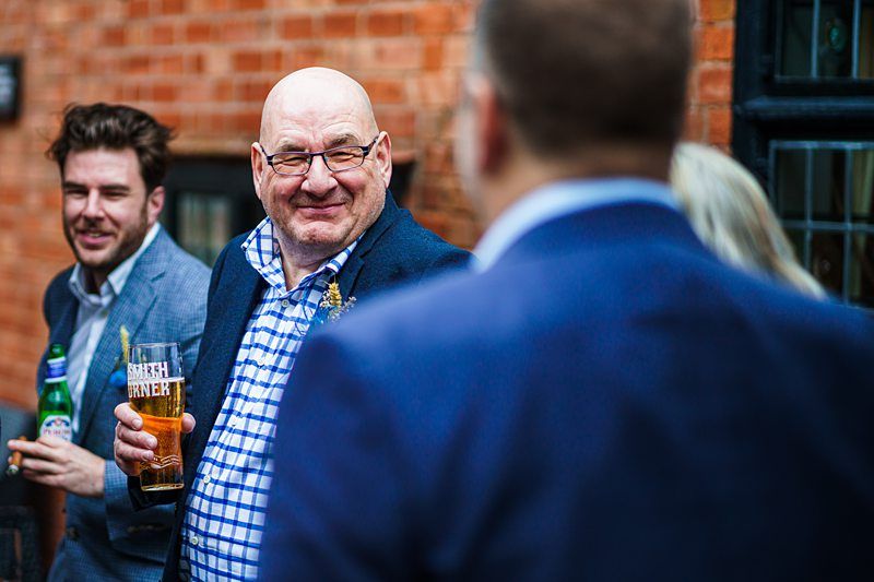 A bald man wearing glasses and a blue plaid shirt stands with a beer glass in hand, smiling and conversing with a group outside a brick building. Another man with a beer is partially visible nearby. Image by Documentary Wedding Photographer Lewis Bishop Photography.