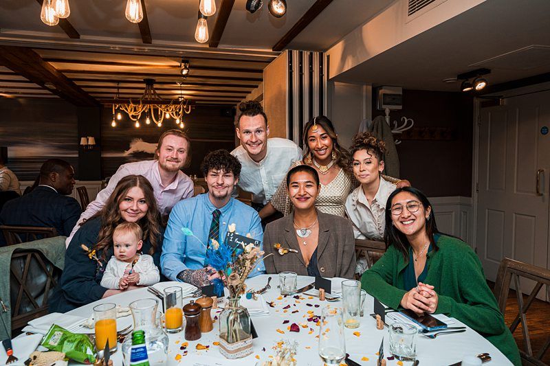 A group of nine people, including one holding a baby, sit around a table in a warmly lit room. Image by Documentary Wedding Photographer Lewis Bishop Photography.
