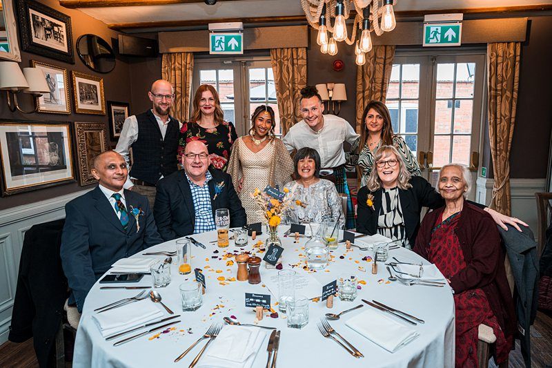 A group of people seated and standing around a round dining table with plates, glasses, and floral decorations in a warmly lit room with framed pictures on the walls. Image by Documentary Wedding Photographer Lewis Bishop Photography.