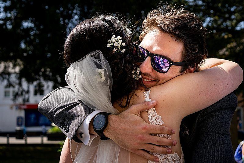 A bride wearing a veil adorned with small flowers embraces a man wearing sunglasses with reflective lenses. The man's smartwatch is visible on his wrist. The scene appears to be outdoors with greenery and buildings in the background. Image by Documentary Wedding Photographer Lewis Bishop Photography.