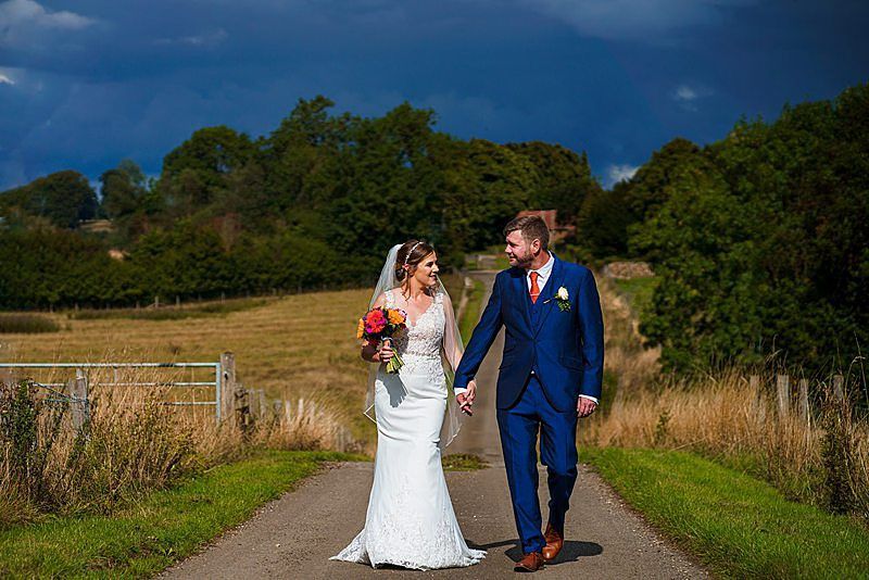 A bride and groom hold hands and walk down a country path with green fields and trees in the background. The bride wears a white dress and veil, holding a colorful bouquet. The groom is in a navy suit with an orange tie. Dark storm clouds loom above them. Image by Documentary Wedding Photographer Lewis Bishop Photography.