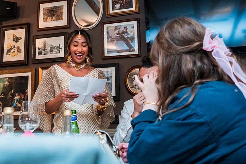 A woman dressed in traditional attire reads from a paper, smiling, while two seated people listen, one covering their face. The background features framed pictures and mirrors on the wall. Image by Documentary Wedding Photographer Lewis Bishop Photography.