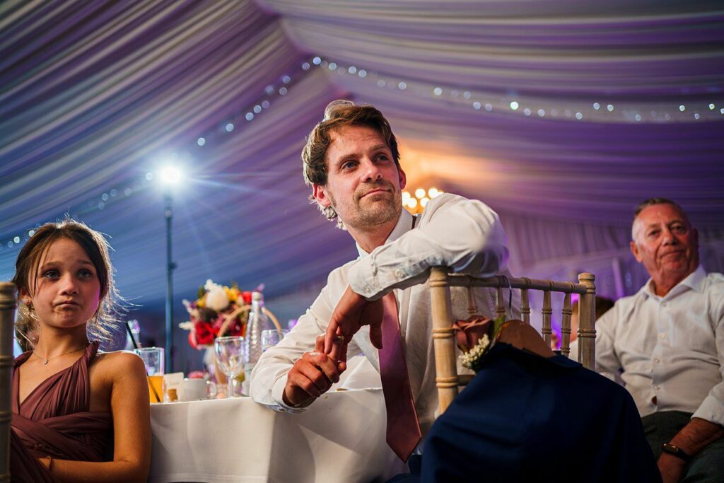 A man in formal attire leans on the back of a chair, accompanied by a young girl in a brown dress, both looking ahead attentively in a decorated and well-lit event tent. An older man in a white shirt sits at the table behind them. Image by Documentary Wedding Photographer Lewis Bishop Photography.