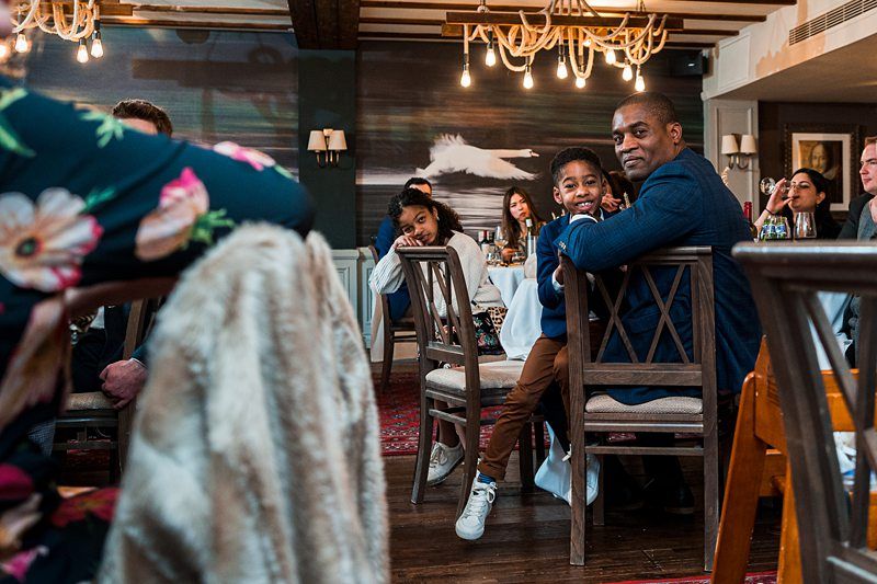 A man and two children sit at a table in a restaurant, smiling at the camera. Other diners are seated at nearby tables. Chandeliers and a wall mural can be seen in the background. Image by Documentary Wedding Photographer Lewis Bishop Photography.