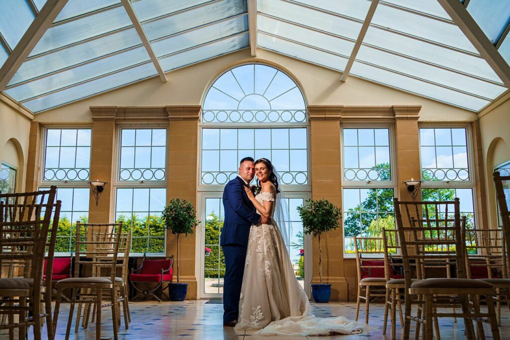 A bride and groom stand together in an elegant, sunlit room with large windows and high ceilings. The bride wears a white gown, and the groom is dressed in a navy blue suit. Empty wooden chairs are arranged on either side, suggesting a wedding ceremony setting. Image by Documentary Wedding Photographer Lewis Bishop Photography.
