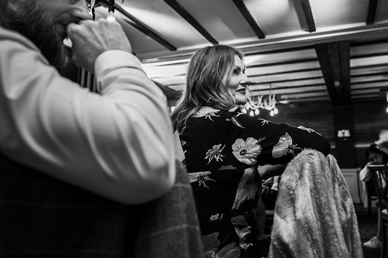 A woman in a floral top and a man in a white shirt with a beard sit in a dimly lit room with exposed wooden beams. The woman is smiling and looking to the side. Image by Documentary Wedding Photographer Lewis Bishop Photography.