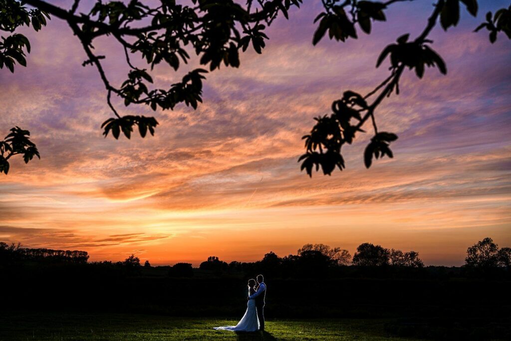 A couple dressed in wedding attire stands lovingly together in an open field at sunset, with vibrant orange, pink, and purple hues filling the sky. Silhouettes of trees and branches frame the serene, romantic scene. Image by Documentary Wedding Photographer Lewis Bishop Photography.