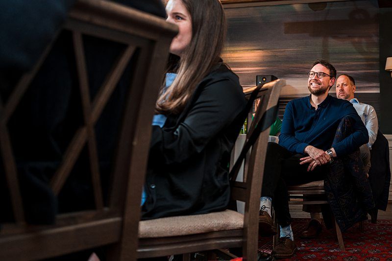 People sitting in wooden chairs, some smiling and attentively listening during an indoor event. Image by Documentary Wedding Photographer Lewis Bishop Photography.