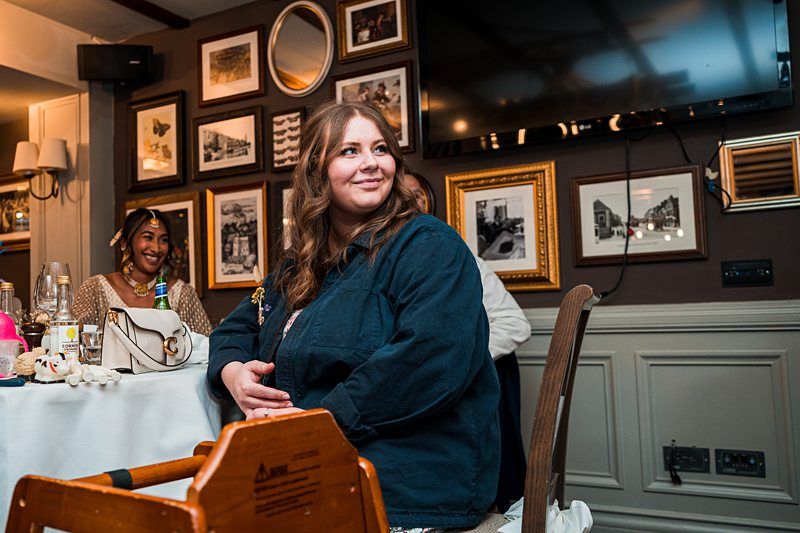 A woman sits at a table, smiling and looking off to the side. The room has framed pictures on the wall and a television. Other individuals and table settings are partially visible in the background. Image by Documentary Wedding Photographer Lewis Bishop Photography.