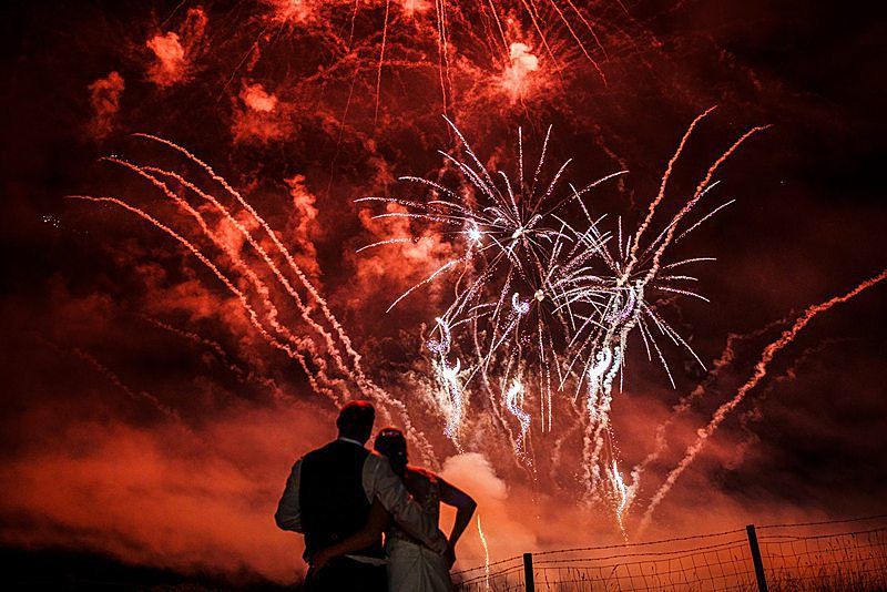 A couple stands with their backs to the camera, watching a night sky filled with bright, colorful fireworks. The sky is illuminated with bursts of red and white, and smoke trails add to the vibrant display. A fence is visible in the foreground. Image by Documentary Wedding Photographer Lewis Bishop Photography.