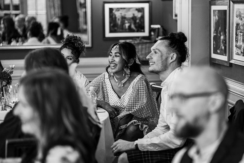 Guests seated at a table, smiling and laughing during an indoor gathering, with framed artwork on the wall in the background. Image by Documentary Wedding Photographer Lewis Bishop Photography.