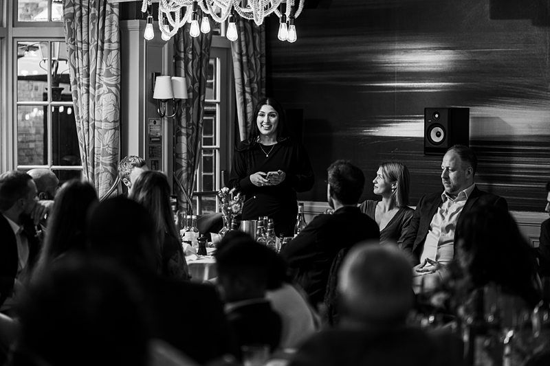 A woman stands and speaks to a seated audience at a formal indoor event, with a chandelier hanging overhead and patterned curtains in the background. Black and white photo. Image by Documentary Wedding Photographer Lewis Bishop Photography.