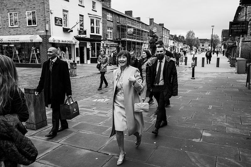 A group of people, dressed in business attire, walk down a stone-paved street lined with buildings and shops on a cloudy day. Image by Documentary Wedding Photographer Lewis Bishop Photography.