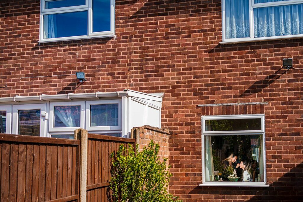 A brick house with two upper windows and a lower window, where a person is visible looking outside. The house has a small attached white structure and a wooden fence with some greenery in the foreground. The scene is bathed in sunlight. Image by Documentary Wedding Photographer Lewis Bishop Photography.