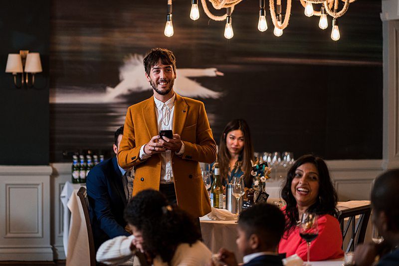 A man in a brown blazer stands at a formal gathering with a phone in hand, smiling. People are seated around tables, and a chandelier with exposed bulbs hangs above. Image by Documentary Wedding Photographer Lewis Bishop Photography.