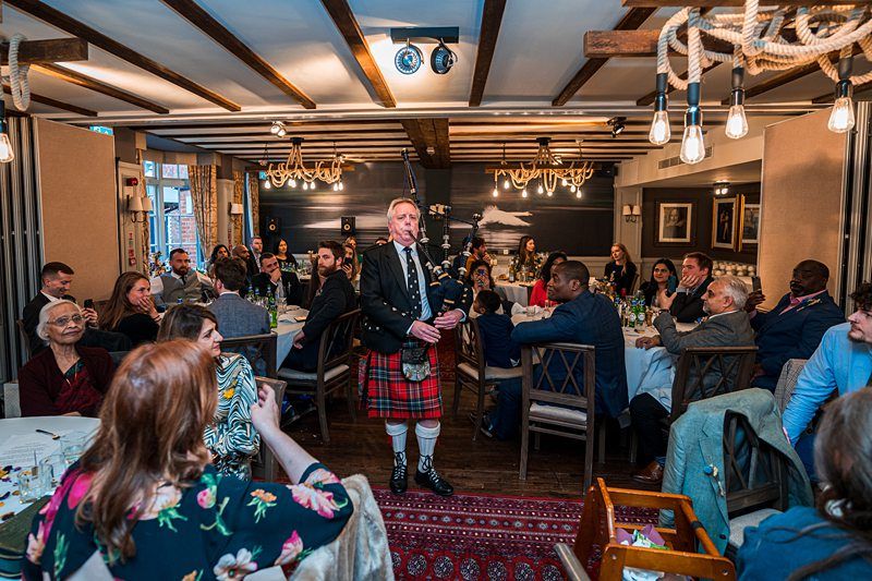 A bagpiper in traditional Scottish attire performs in a warmly lit, rustic dining room filled with seated guests. Tables are adorned with white tablecloths and various plates and glasses. Image by Documentary Wedding Photographer Lewis Bishop Photography.