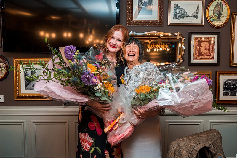 Two women standing close together, smiling, and holding large bouquets of flowers. They are in a room decorated with framed pictures and a mirror on the wall. Image by Documentary Wedding Photographer Lewis Bishop Photography.