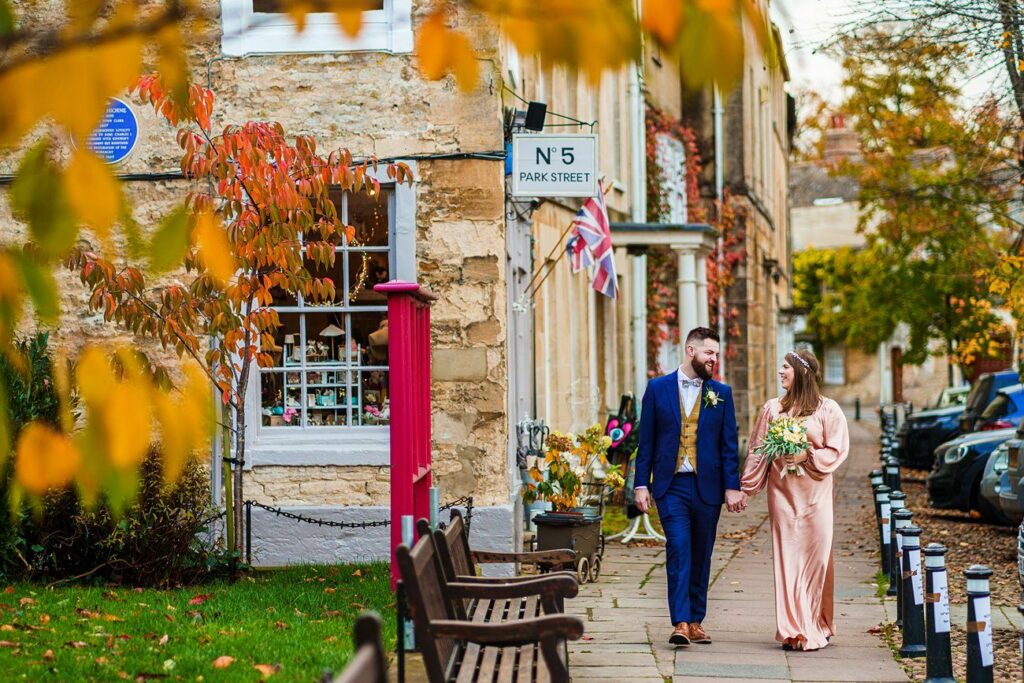 A couple, dressed in formal wedding attire, walk hand in hand along a charming street lined with benches and autumn foliage. The groom wears a navy suit while the bride wears a flowing pink dress and holds a bouquet. They smile at each other, enjoying the moment. Image by Documentary Wedding Photographer Lewis Bishop Photography.