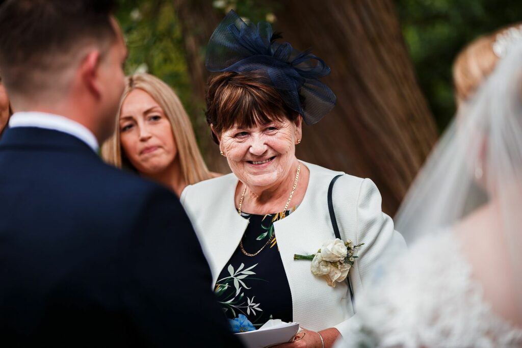 An elderly woman in a white outfit with a floral brooch and navy fascinator smiles warmly at a wedding reception. Two other people, including a bride in a white dress with a veil, are visible in the foreground. Trees are visible in the background. Image by Documentary Wedding Photographer Lewis Bishop Photography.
