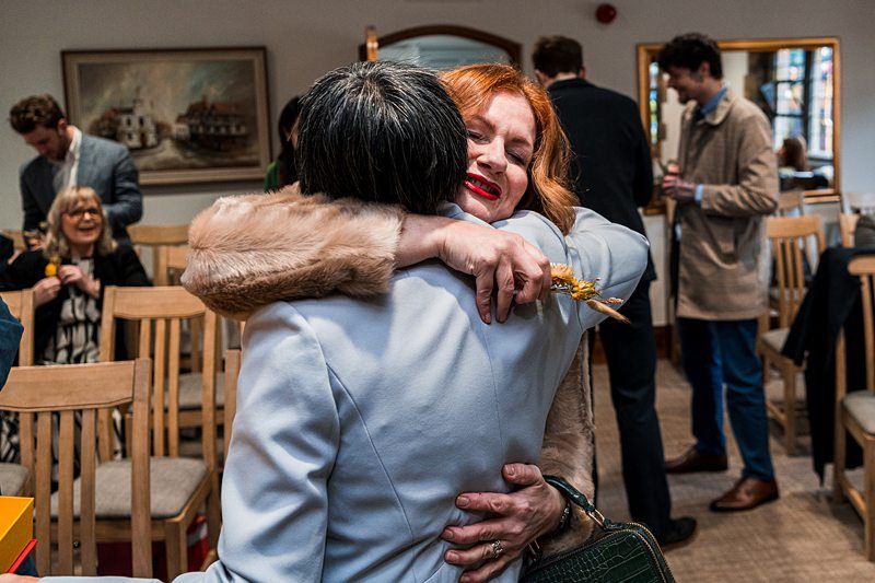 A woman with red hair and a colorful scarf hugs another woman with short gray hair at a social gathering in a room with wooden chairs and paintings on the wall. Other people in the background are smiling and socializing. Image by Documentary Wedding Photographer Lewis Bishop Photography.