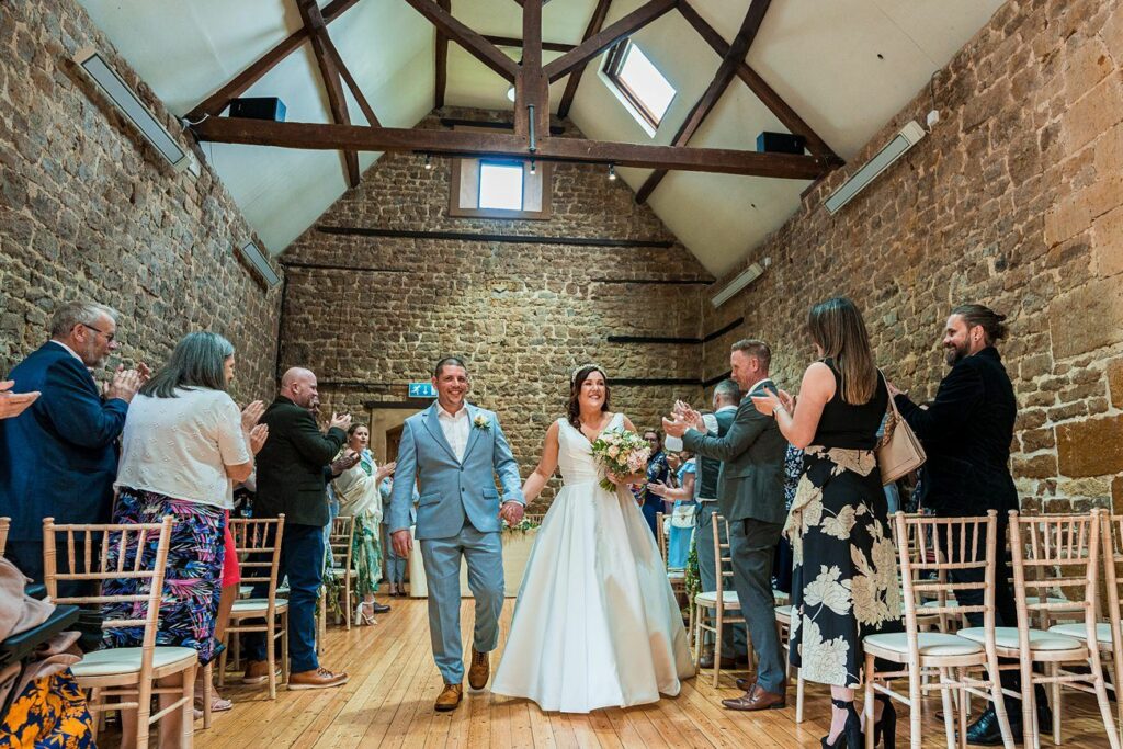 A couple walks down the aisle holding hands in a rustic venue with exposed stone walls and wooden beams, surrounded by applauding guests. The bride holds a bouquet and wears a white gown. Image by Documentary Wedding Photographer Lewis Bishop Photography.